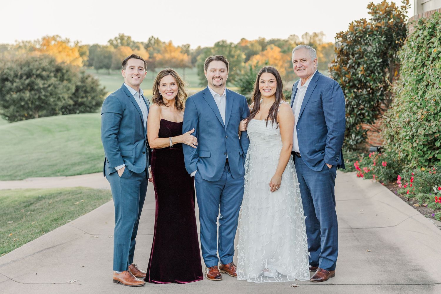A wedding party in blue suits and formal attire poses together outdoors during autumn.