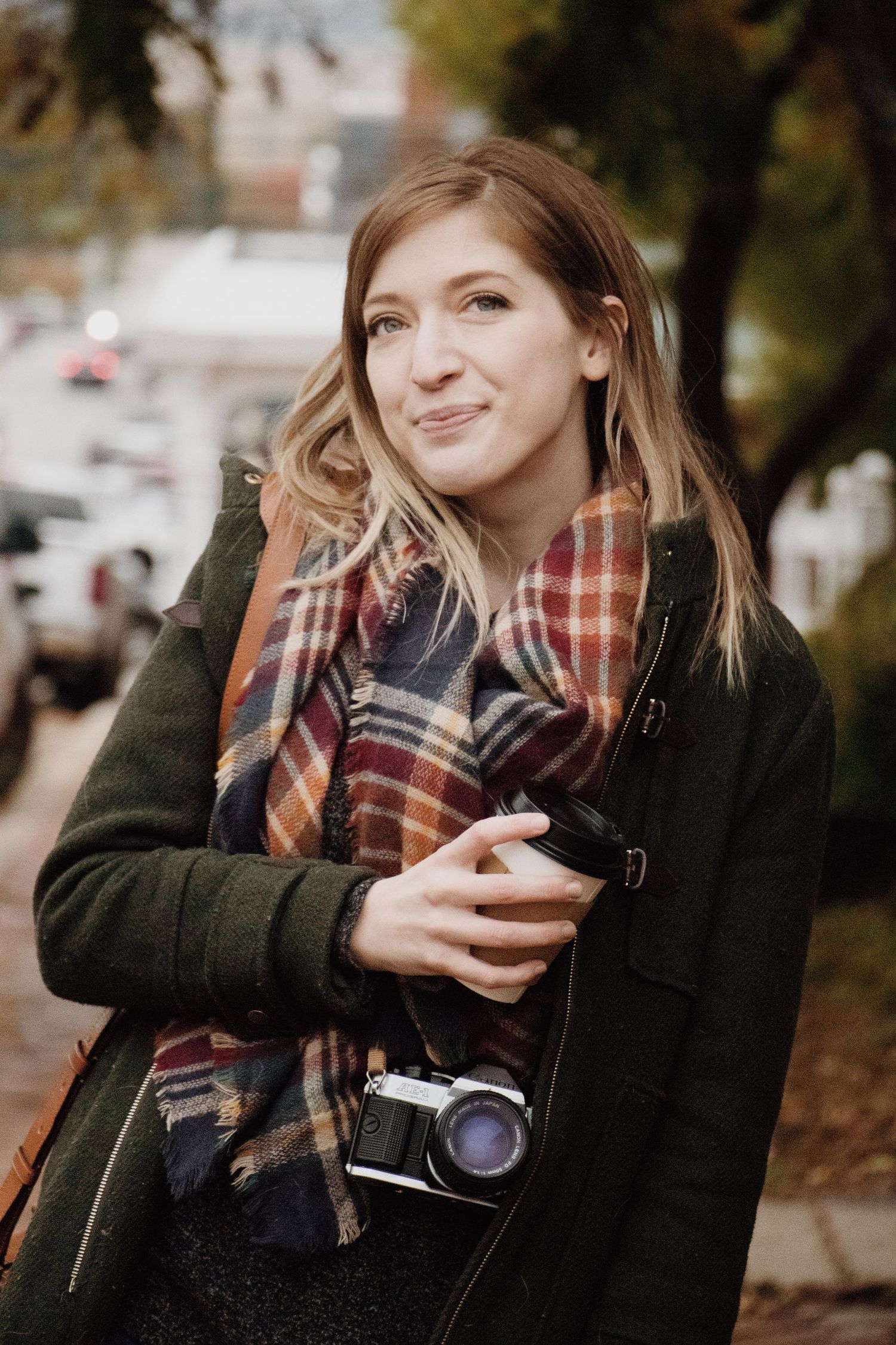 A Kansas City photographer wearing a coat and scarf holds a film camera and cup of coffee as she smiles softly and looks beyond the camera.