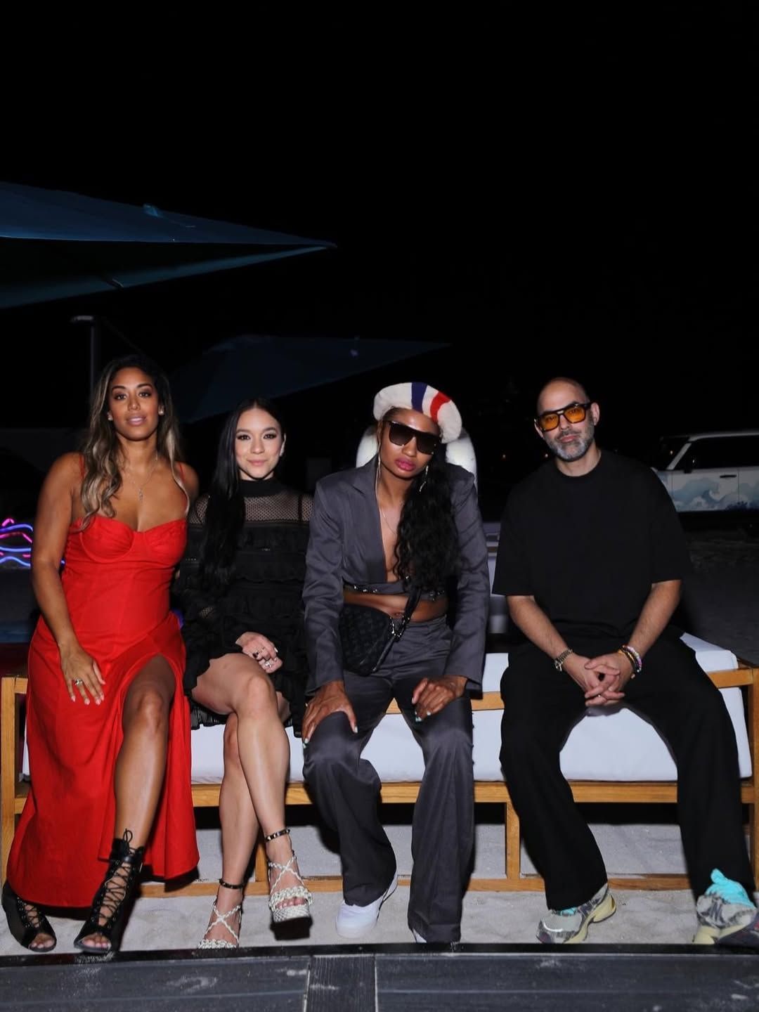 A group of people sit casually together on a wooden bench at night wearing dark clothing and red accents.