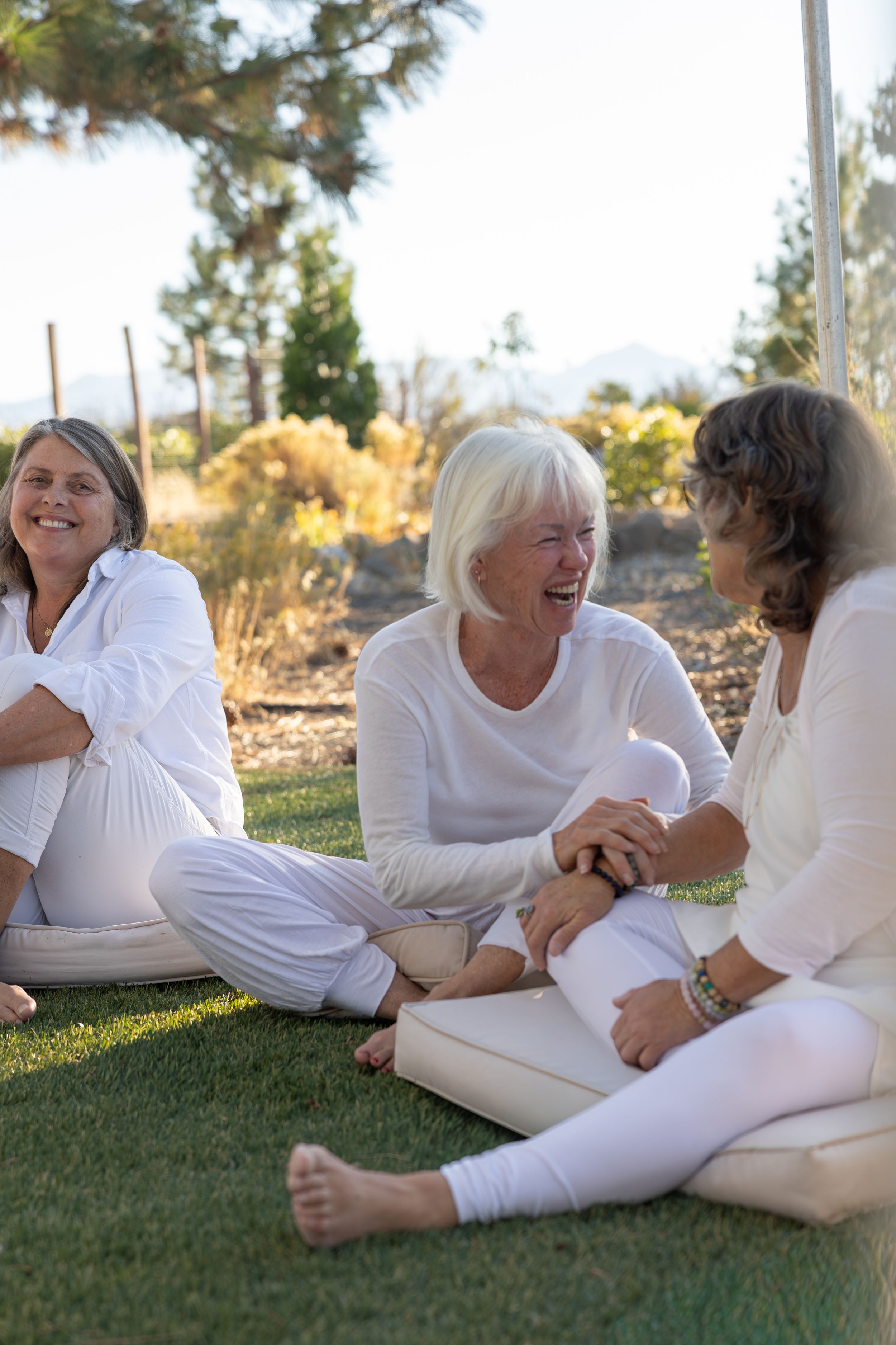 Group of people in white clothing relaxing and laughing together on grass outdoors.