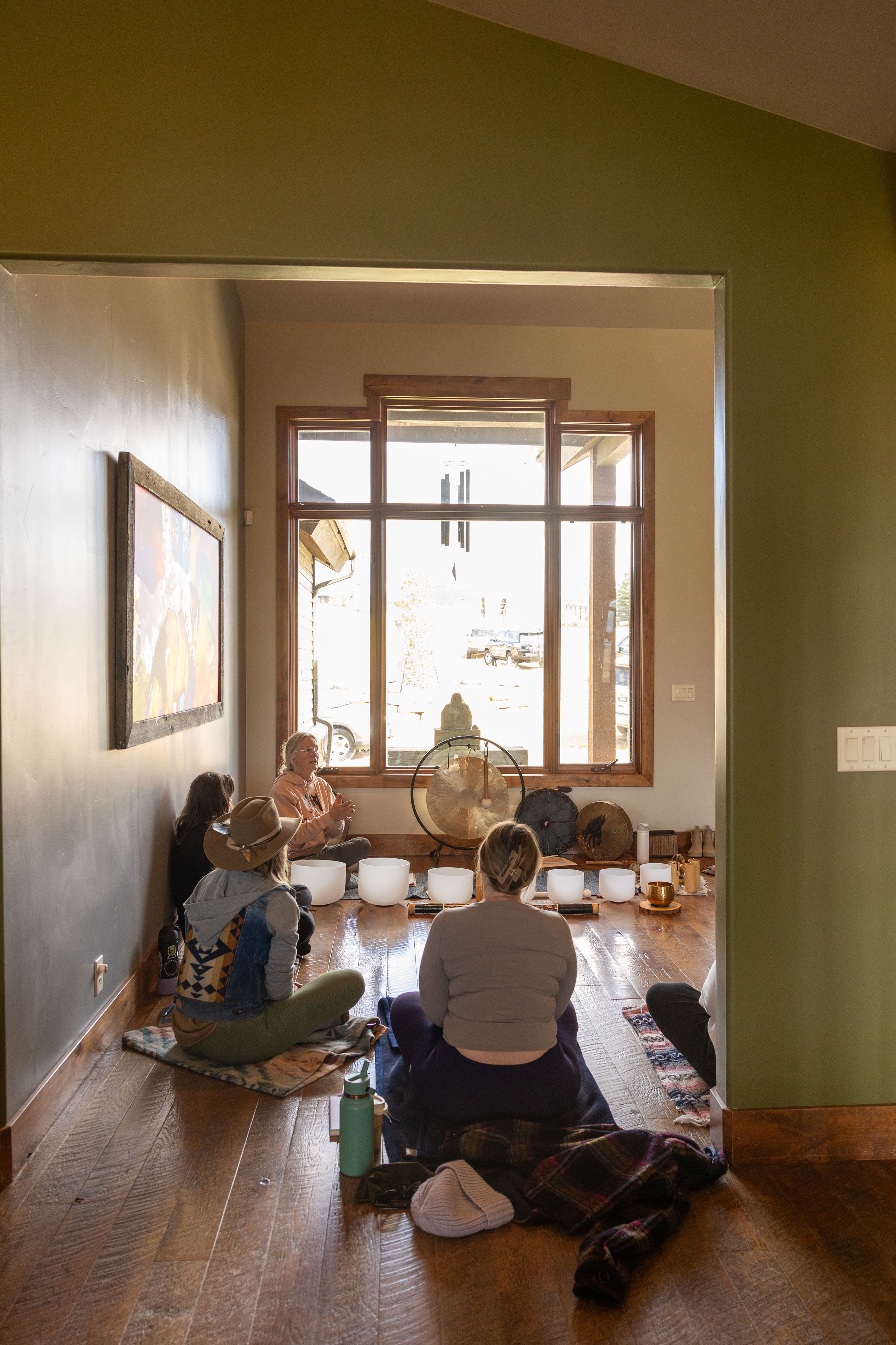 A group of people sit on the floor in a sunlit room with wooden floors and windows, viewing something off-camera.