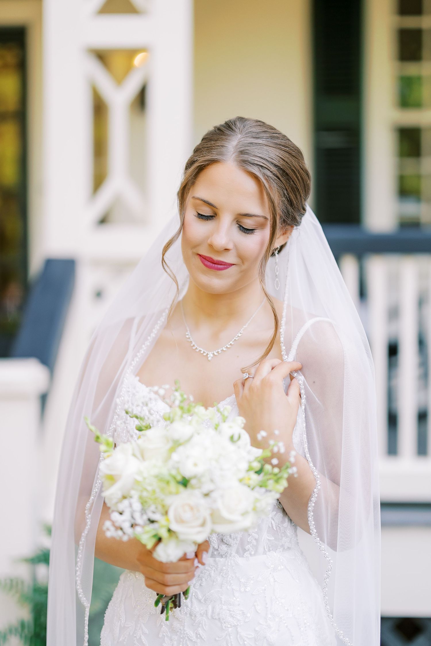 A bride in a white lace dress stands on a covered porch with white columns and black shutters.