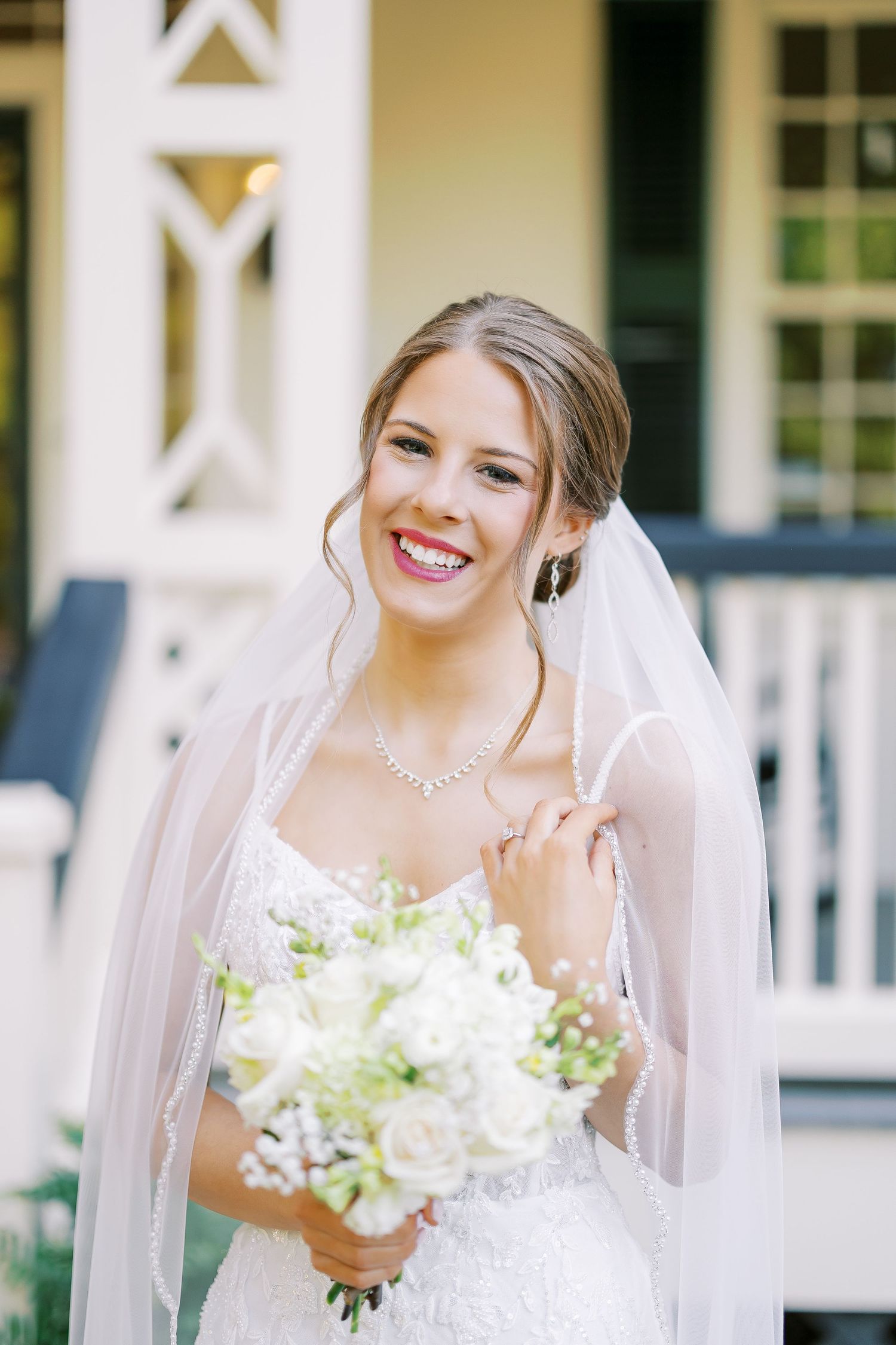A bride in a white wedding dress holds a bouquet of white flowers while wearing a flowing veil on a sunny day.