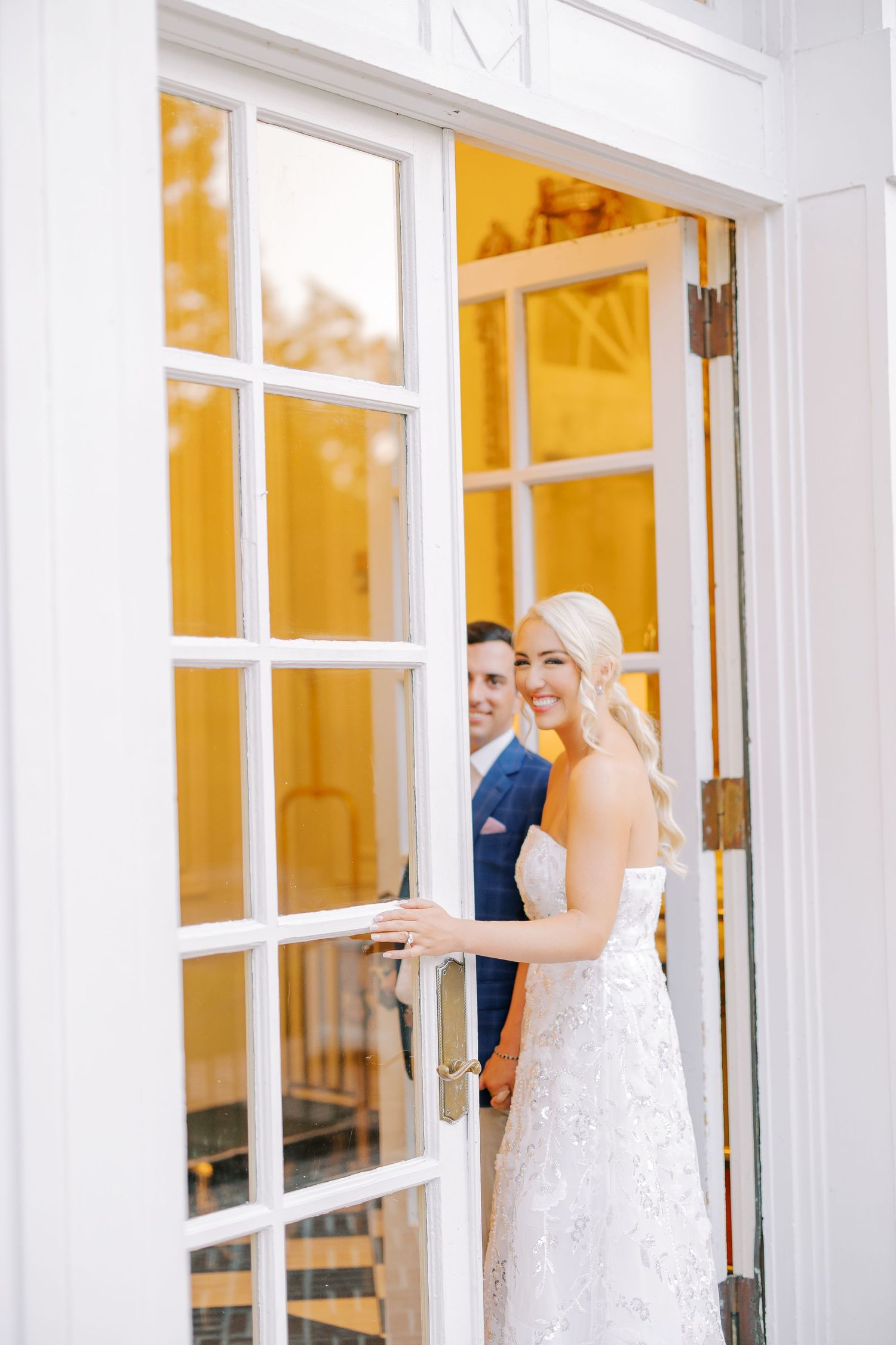 A wedding party in white dresses stands by elegant French doors with golden light streaming through the windows.