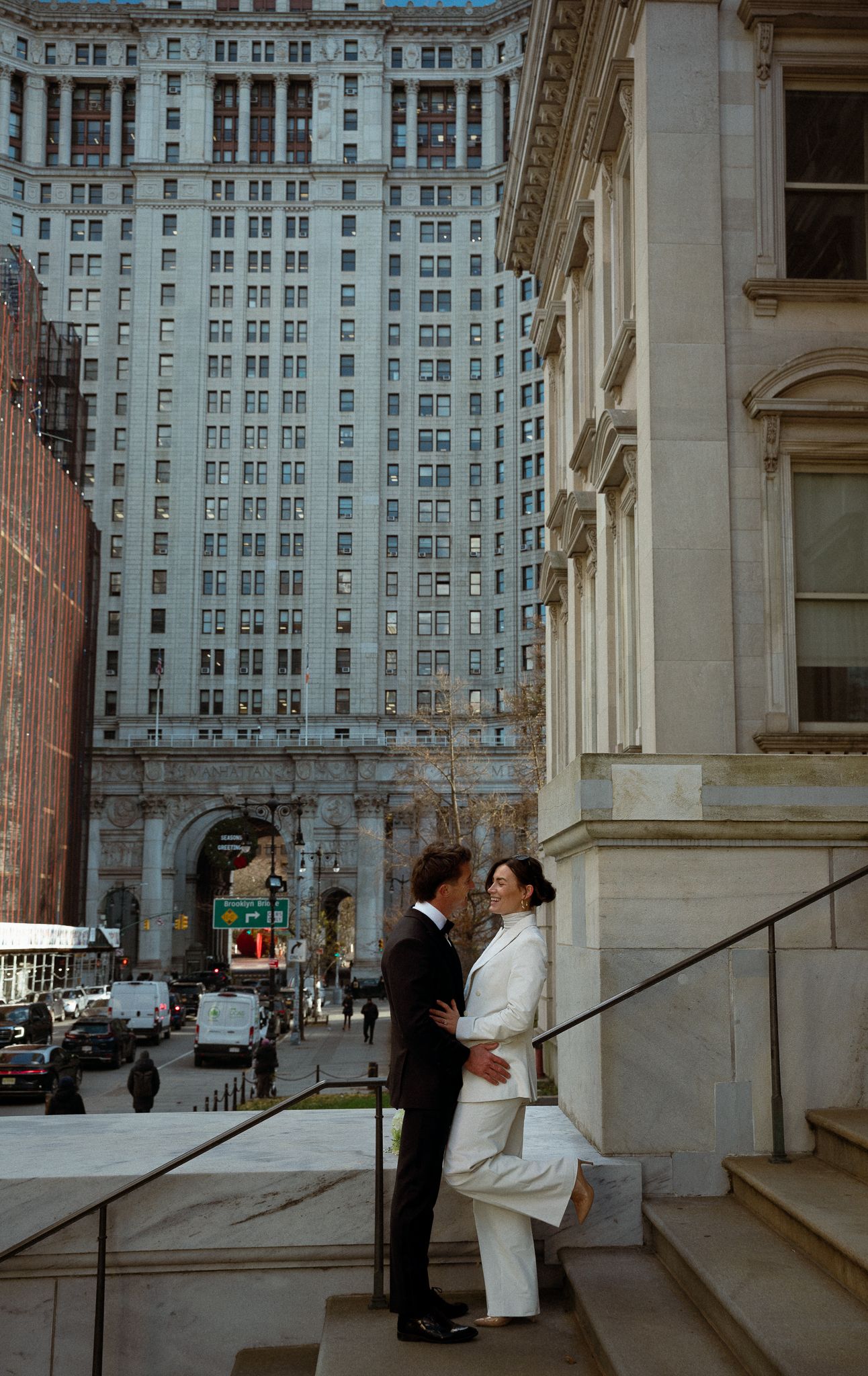 A couple in formal attire stands on the steps of a historic building with city views.