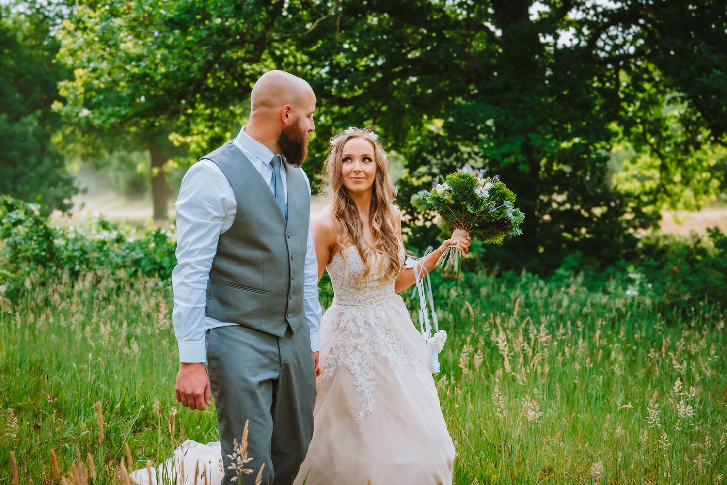 A couple in wedding attire walks together through a lush green meadow on a sunny summer day.