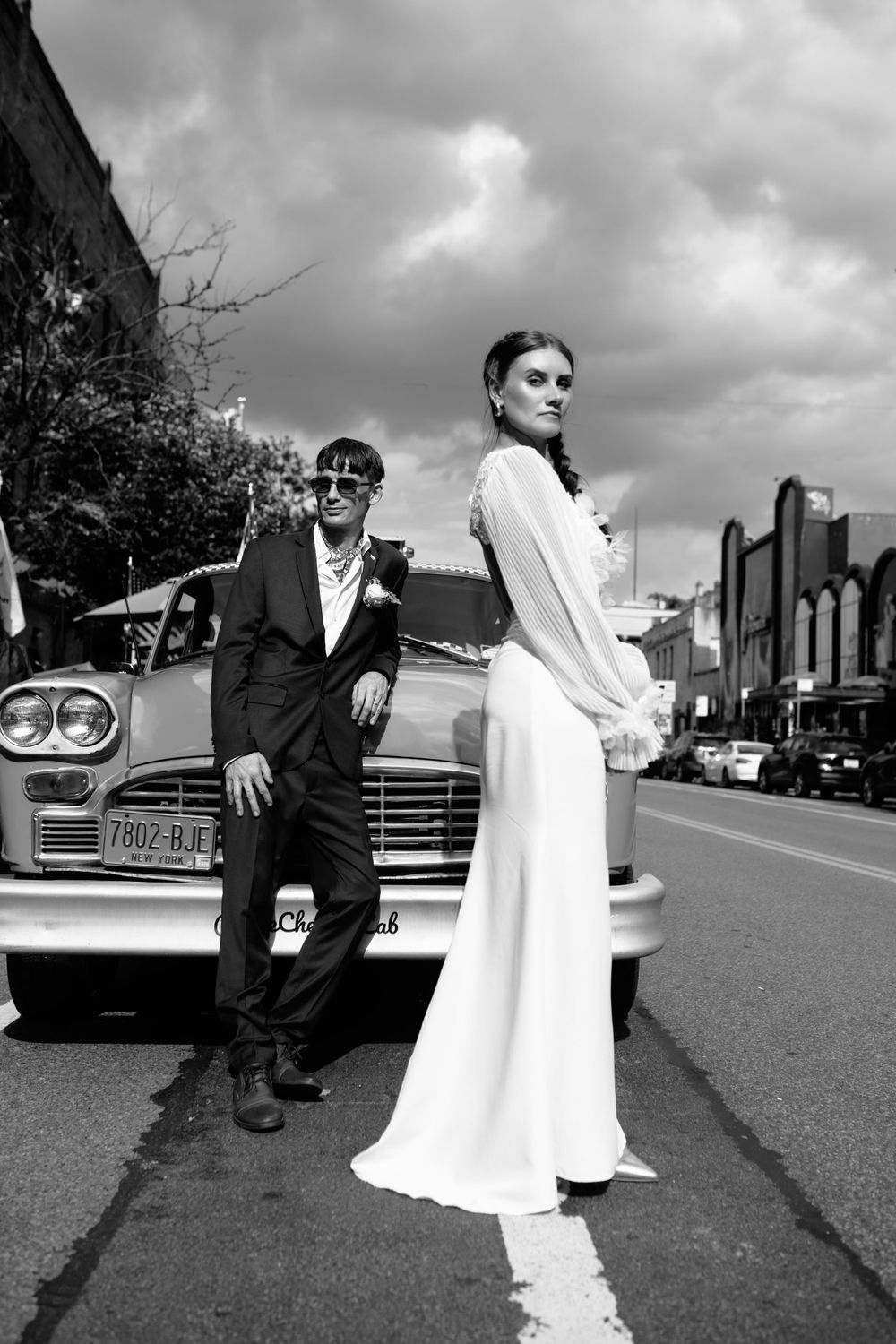 Vintage black and white street photography of a couple posing with a classic car in an urban setting.