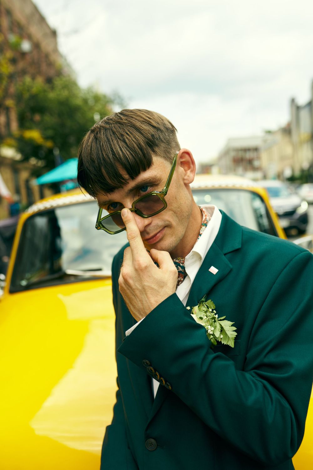Stylish groom in emerald green suit jacket and sunglasses poses beside yellow taxi.