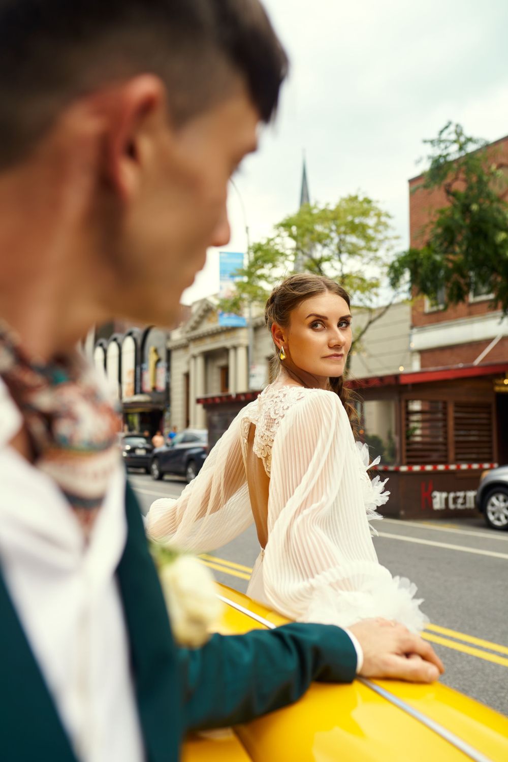 Wedding couple in elegant attire pose dramatically by yellow taxi on city street.