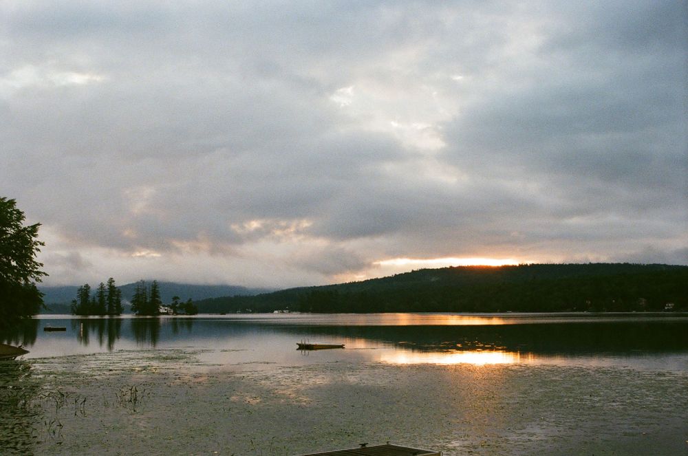 Dramatic sunset over a serene lake with mountains and trees silhouetted against cloudy skies.