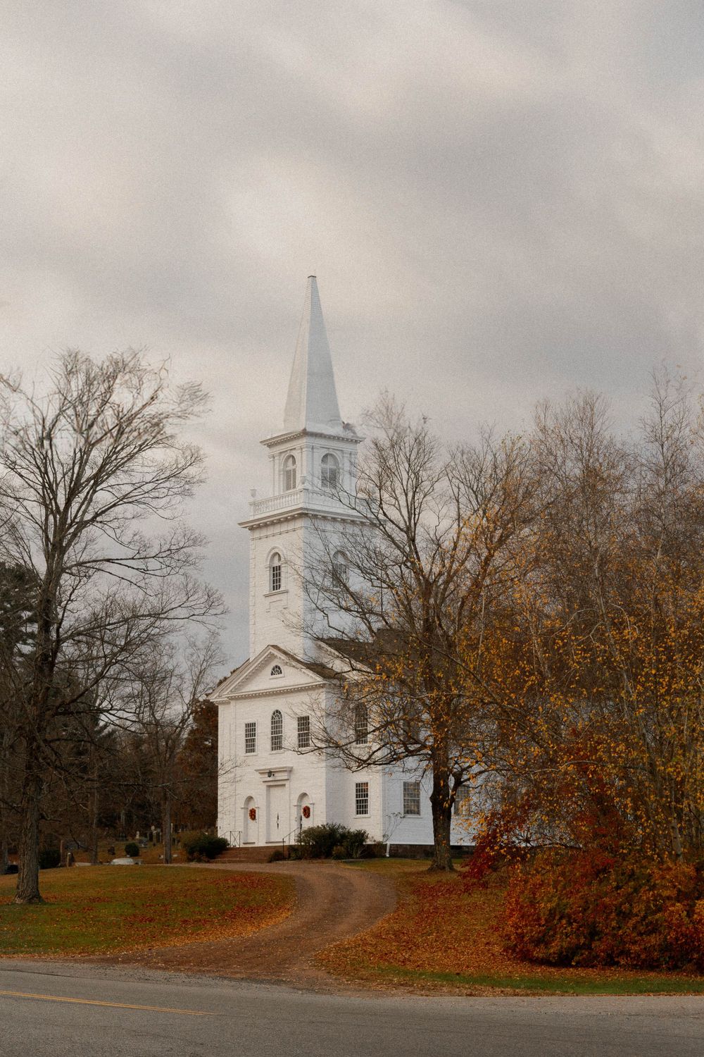 Classic white New England church with tall steeple surrounded by autumn trees on an overcast day.