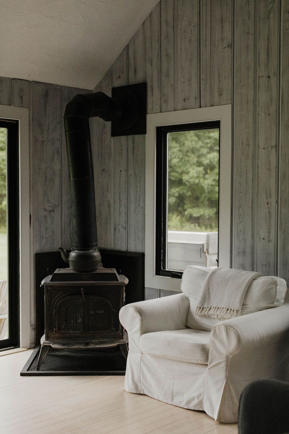 Cozy living room corner with black wood stove and white slipcovered armchair beside windows.