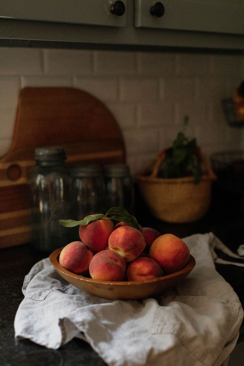 Fresh peaches arranged in wooden bowl on kitchen counter with white cloth.
