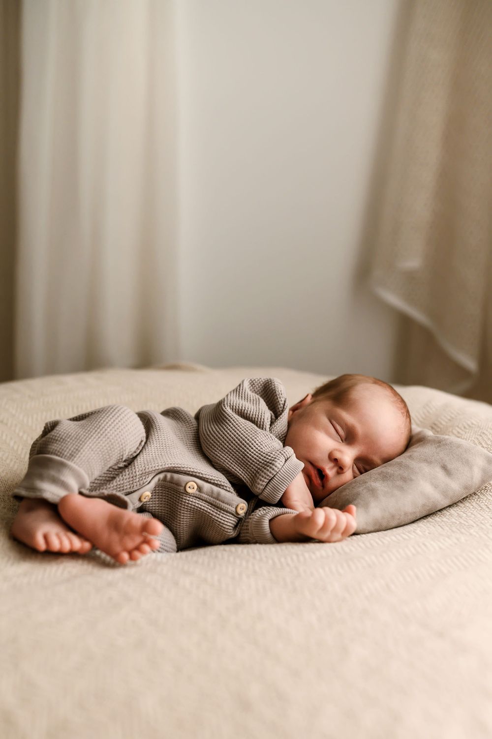 Baby sleeping peacefully on beige bedding wearing a knitted gray sweater outfit.
