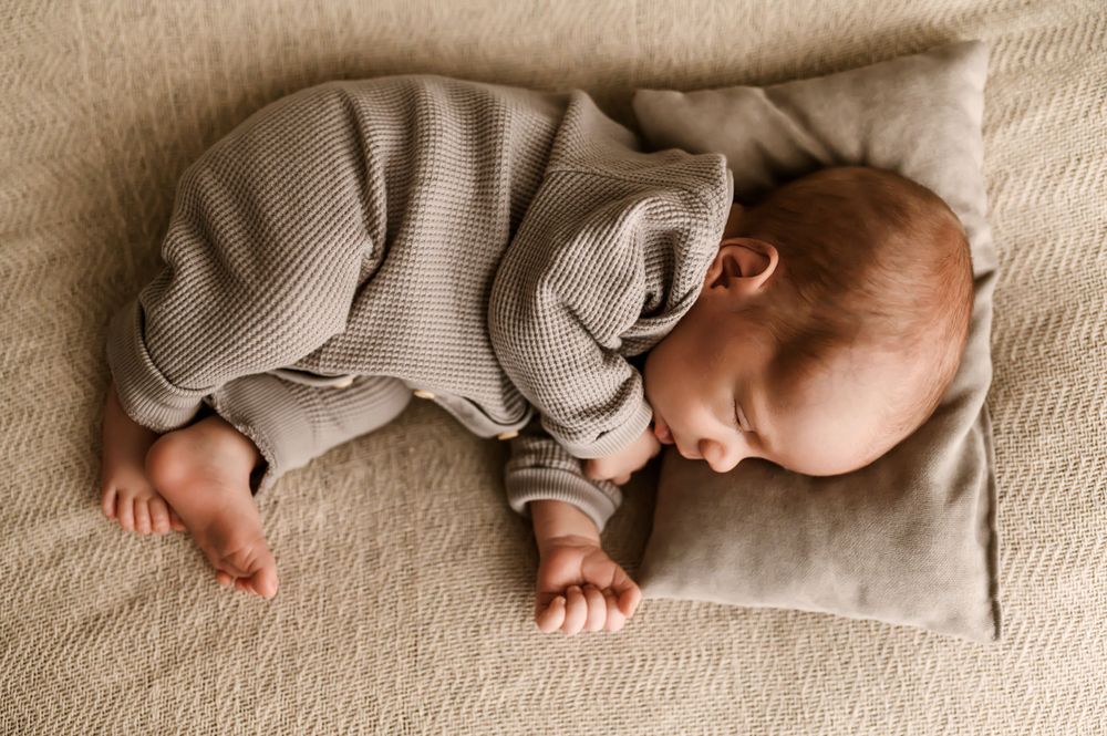 A sleeping baby wearing a cozy gray sweater lies peacefully on a beige cushion.