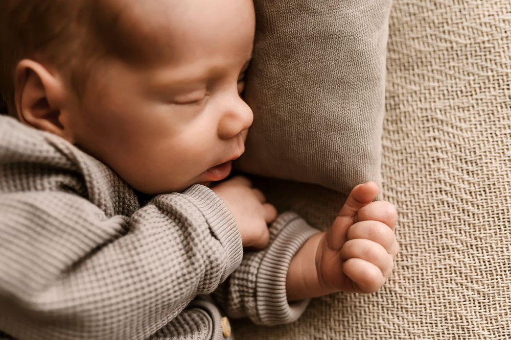 A peaceful sleeping baby in a gray sweater clutches a beige pillow while taking a nap.