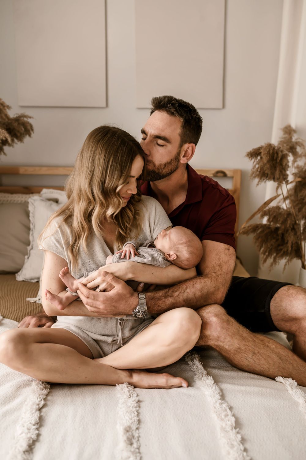 A couple sits intimately on a white bed holding their newborn baby in a soft, natural lit bedroom setting.