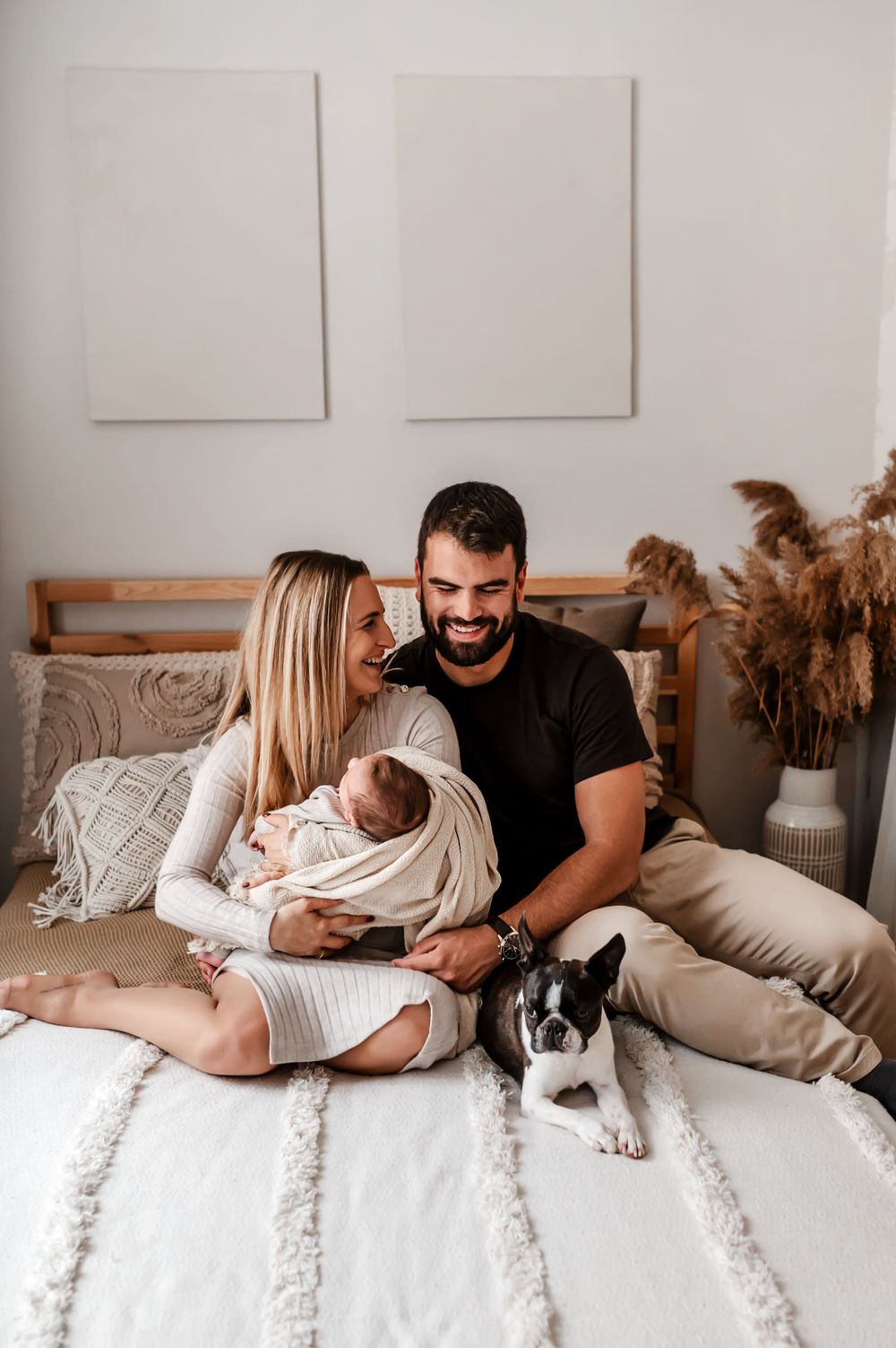 A couple sitting on a white bed with their newborn baby and a small dog in a cozy bedroom setting.