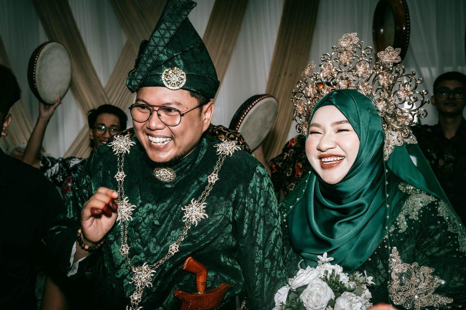 Newlyweds in traditional Malay wedding attire with ornate crowns smile during celebration.