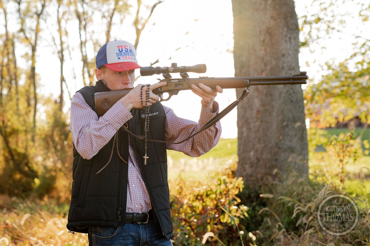 A person in hunting attire aims a rifle in an autumn forest setting.