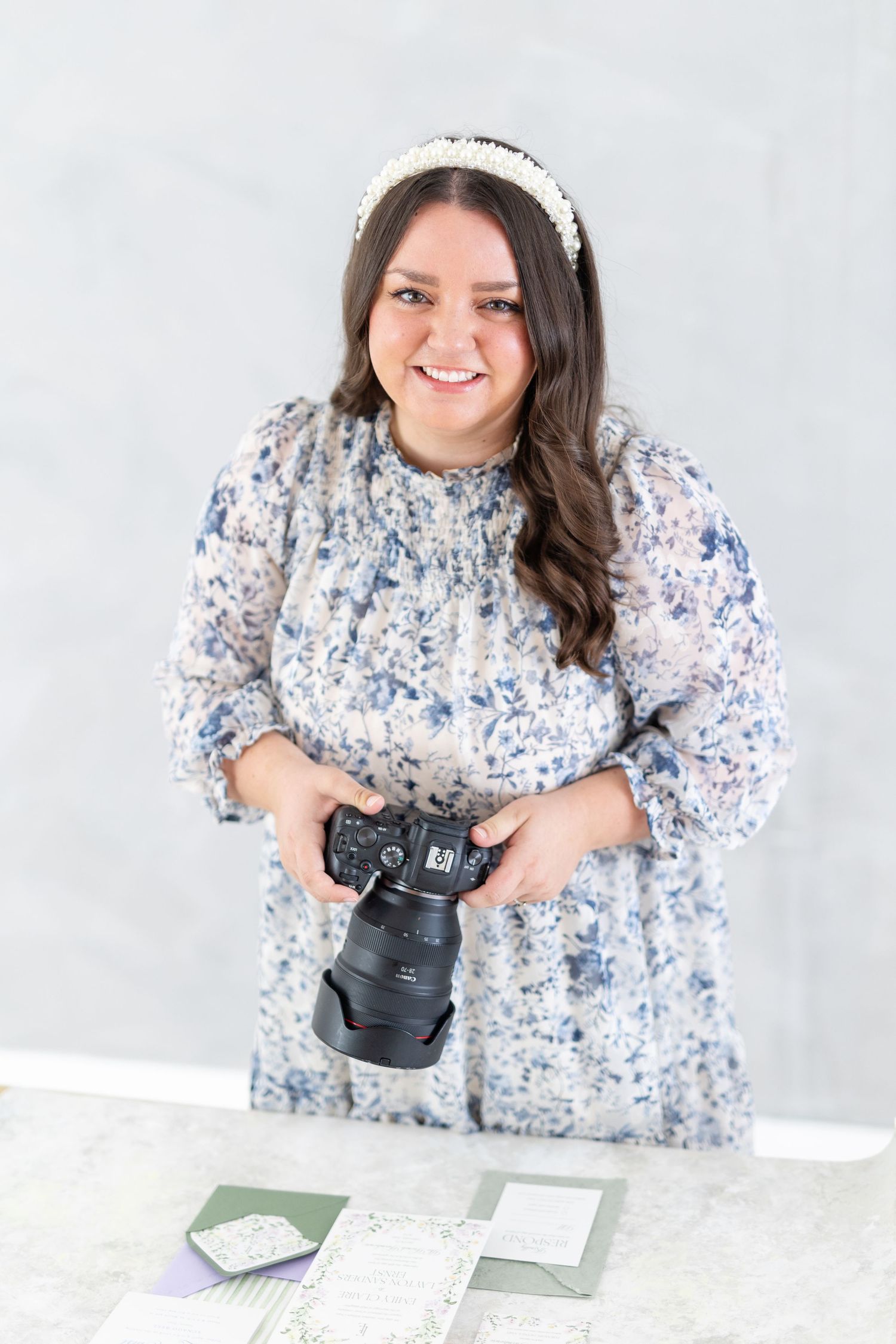 Smiling female photographer in floral dress holding a Canon DSLR camera in a bright studio setting.
