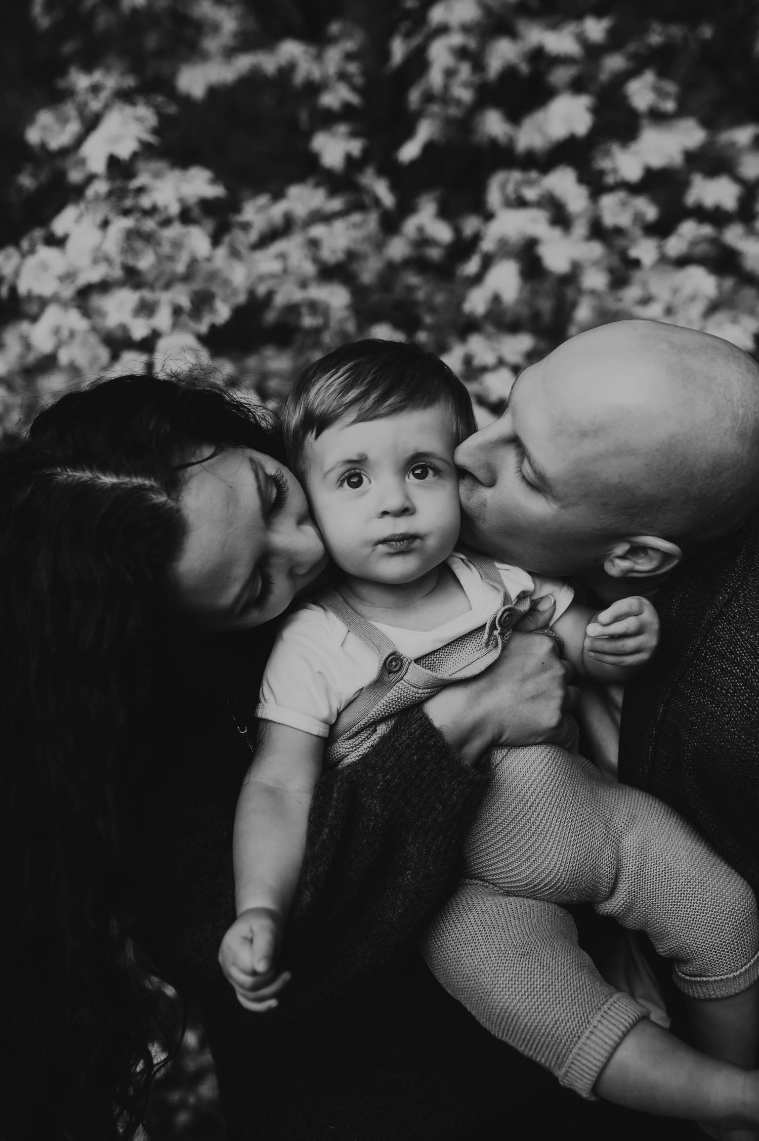 A tender black and white family portrait showing loving kisses being shared with a small child outdoors Grand Rapids Family photographer