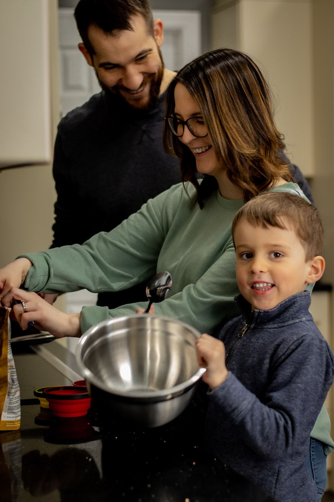 Family cooking together in kitchen with young child helping mix ingredients in large metal bowl.