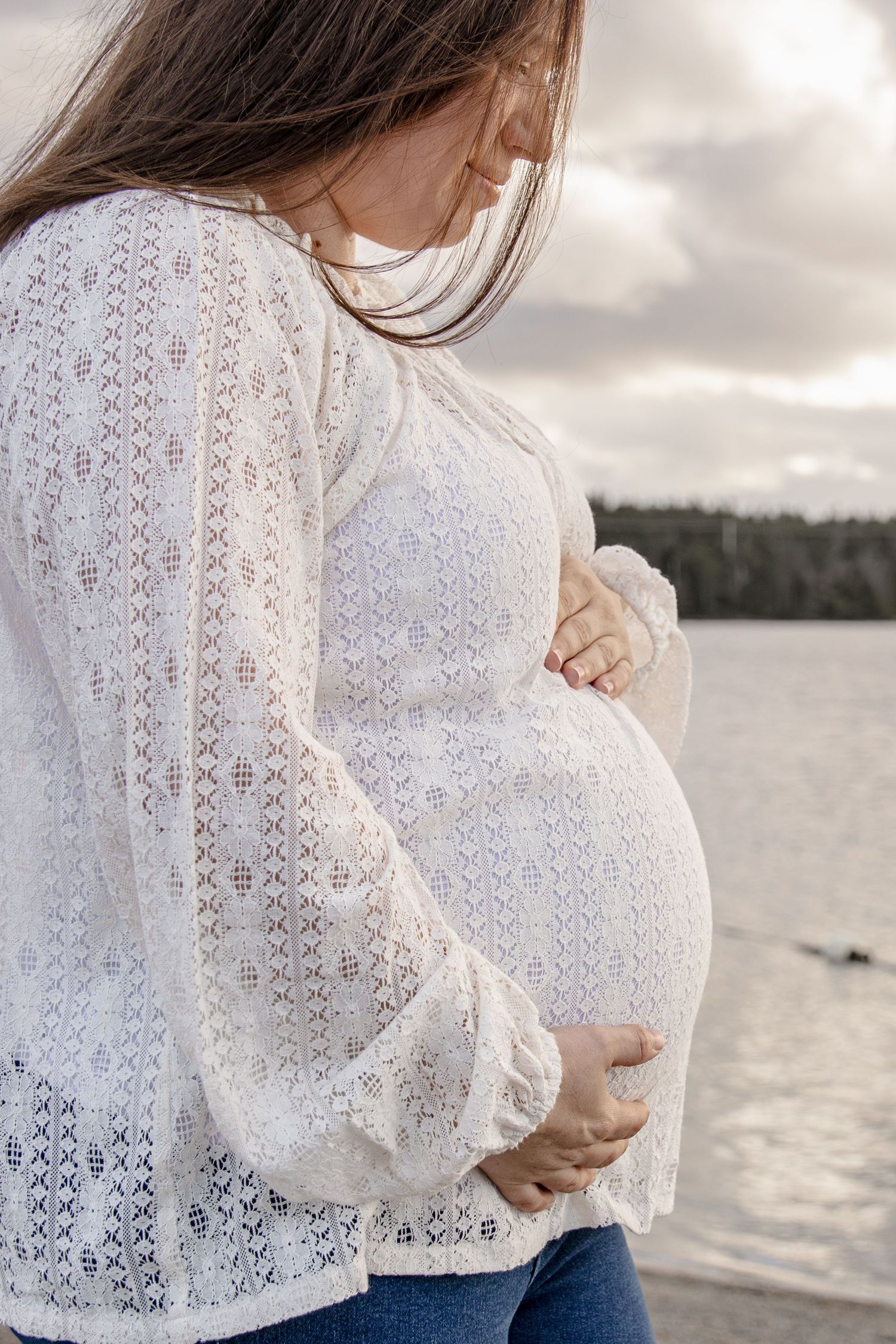 A pregnant person in a white lace blouse stands by a lake, gently cradling their baby bump at sunset.