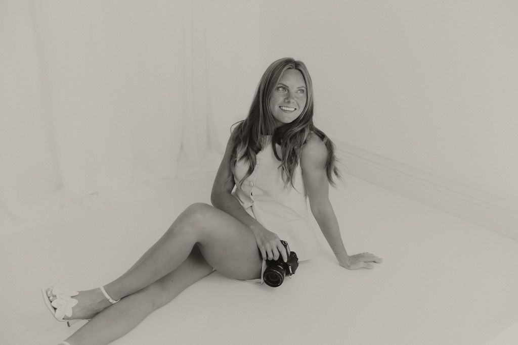 Black and white photo of a smiling woman holding a DSLR camera close to her face with eyes closed.