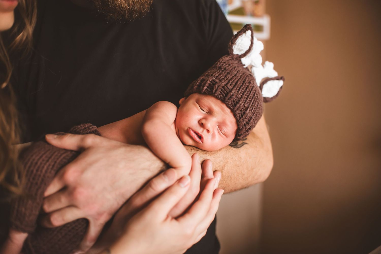 A newborn baby wearing a brown knit hat is gently cradled in someone's hands.