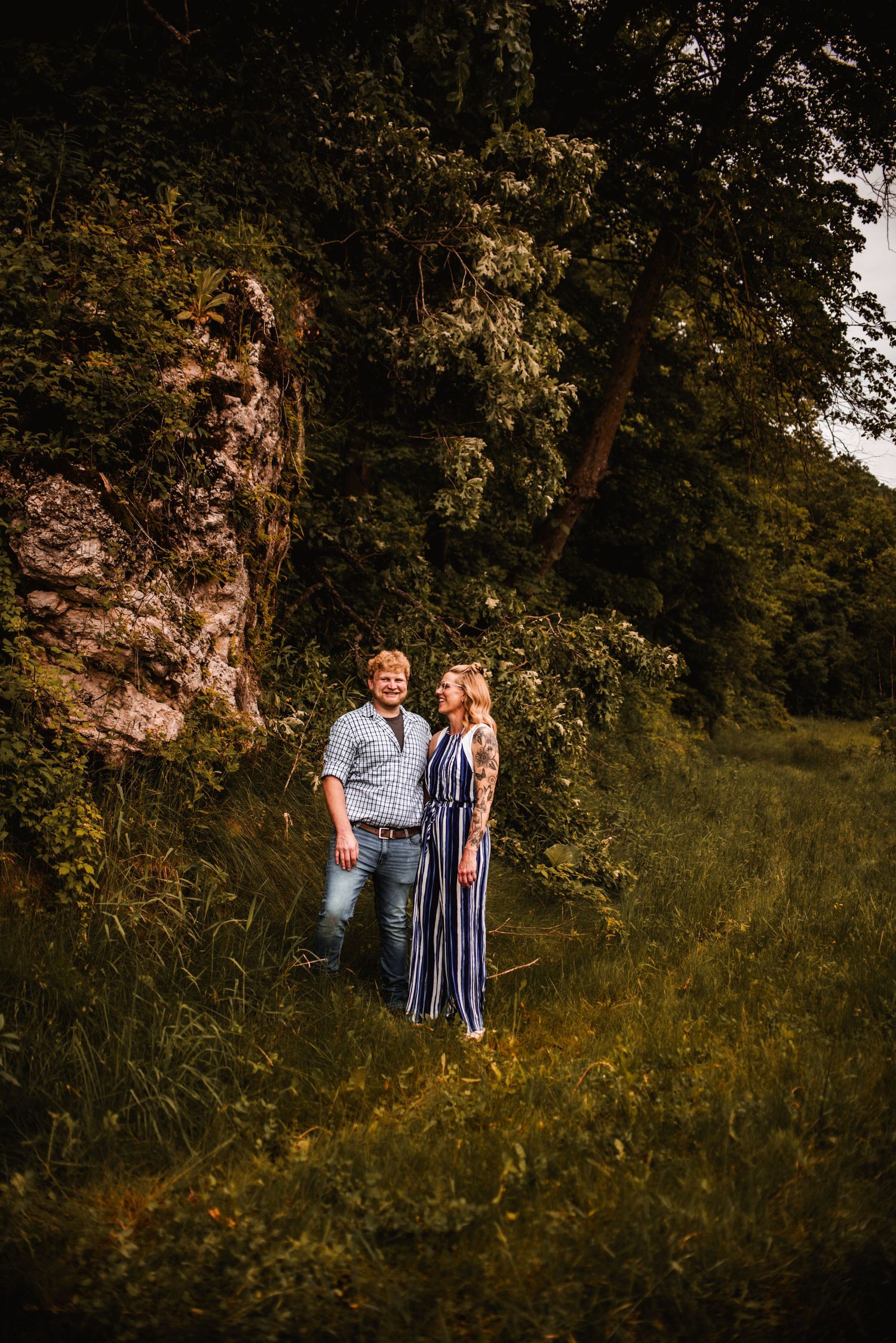 A couple stands together near a rocky cliff face surrounded by lush greenery.