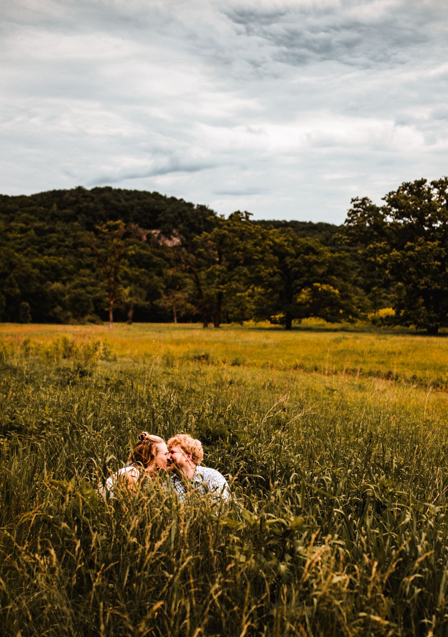 A couple lying in a golden meadow with rolling hills and trees in the background.