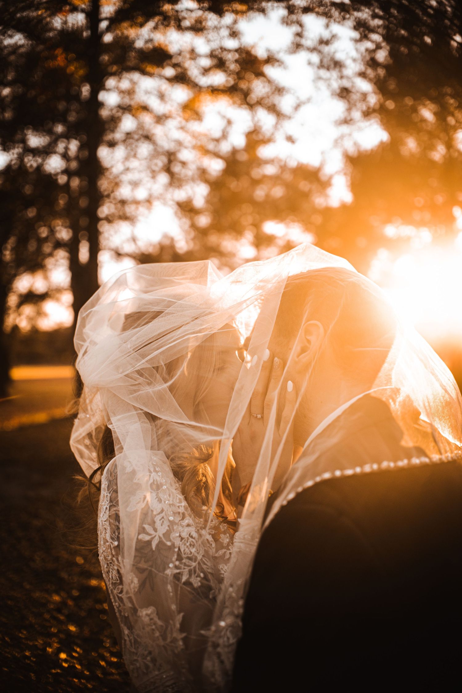 A flowing white wedding veil catches the golden sunset light through trees in a romantic outdoor setting.