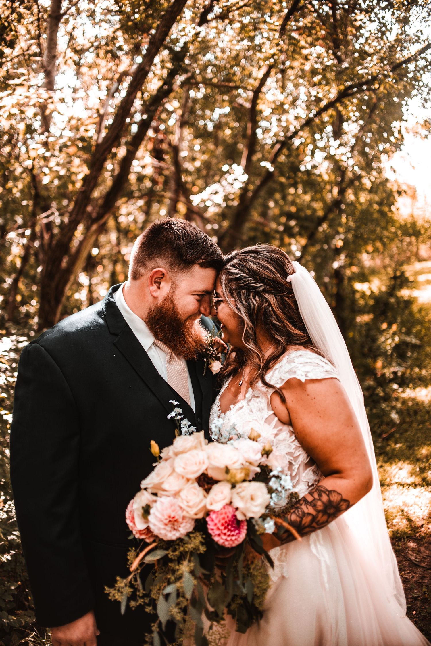 A romantic wedding couple shares an intimate moment in a sunlit wooded setting with a blush pink bouquet.