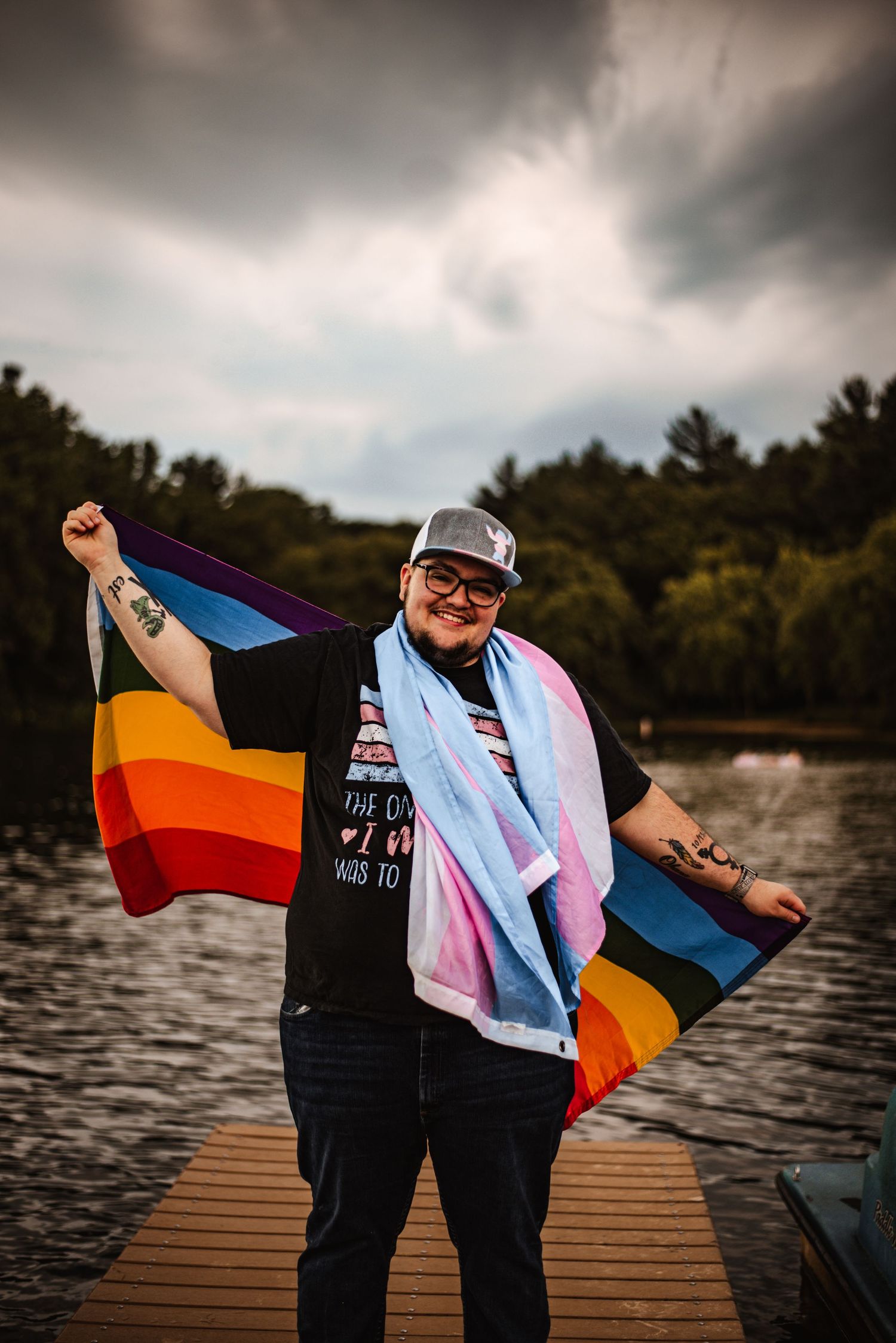 Person on dock displaying pride flags at sunset against a dramatic cloudy sky.