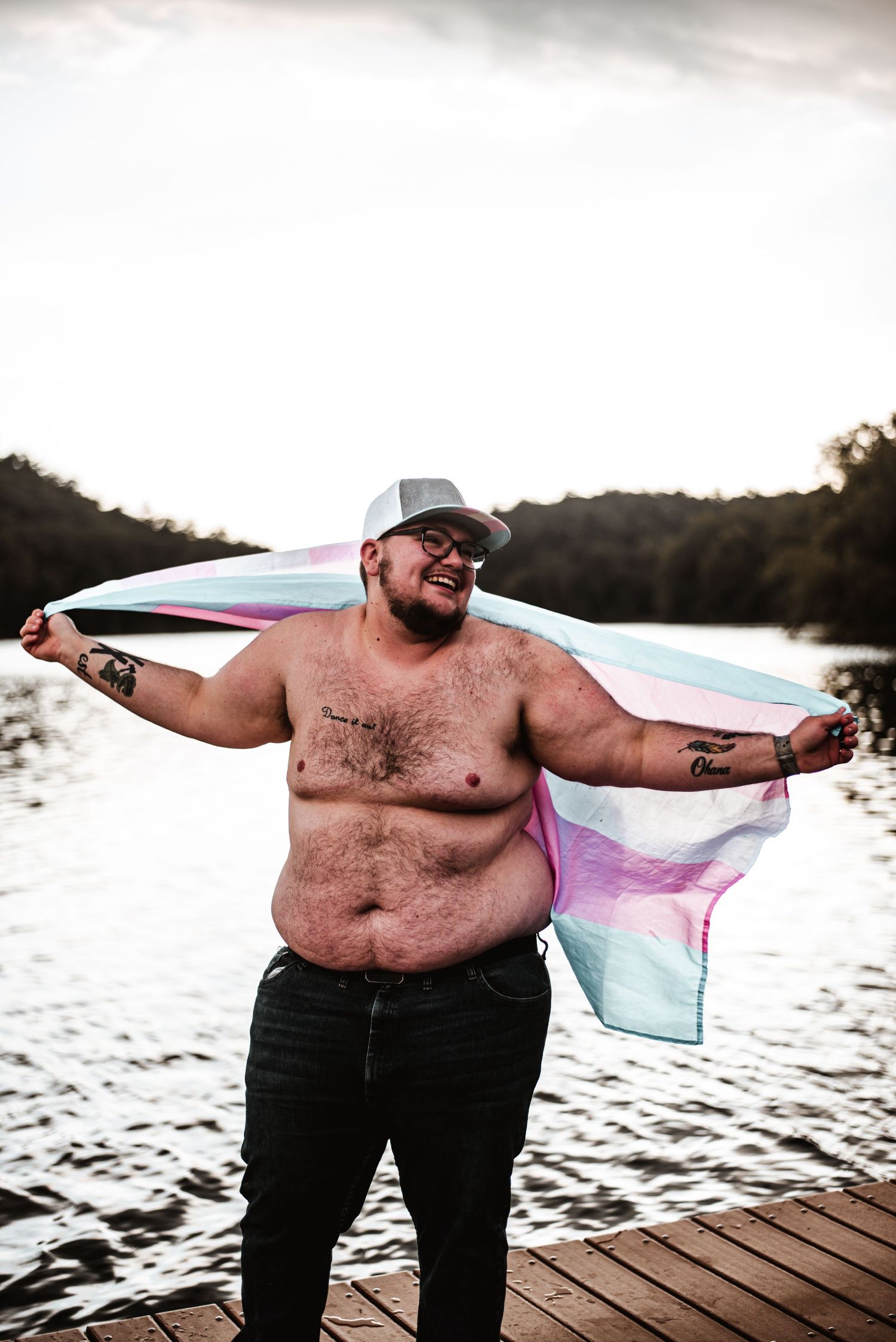Person standing confidently on wooden dock with transgender pride flag over water at sunset.