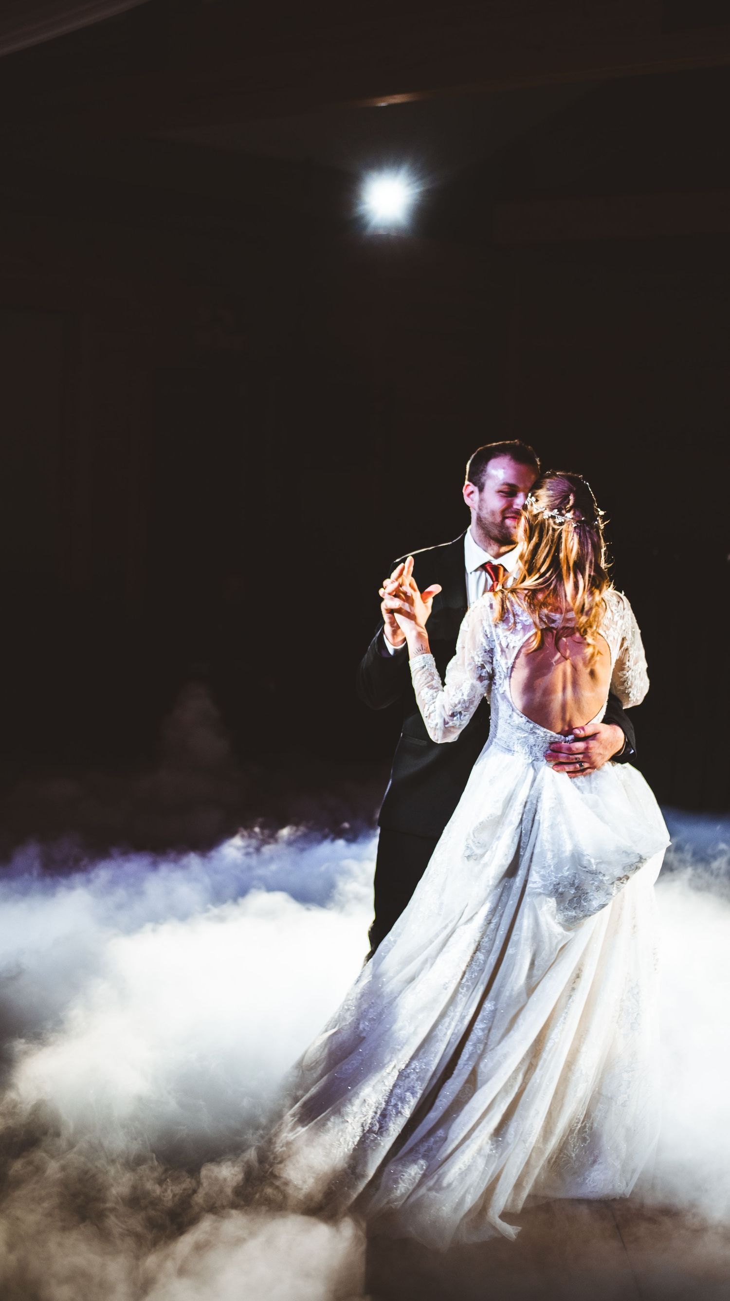 A couple shares their first dance as newlyweds surrounded by romantic fog effects under dramatic lighting at their wedding reception.