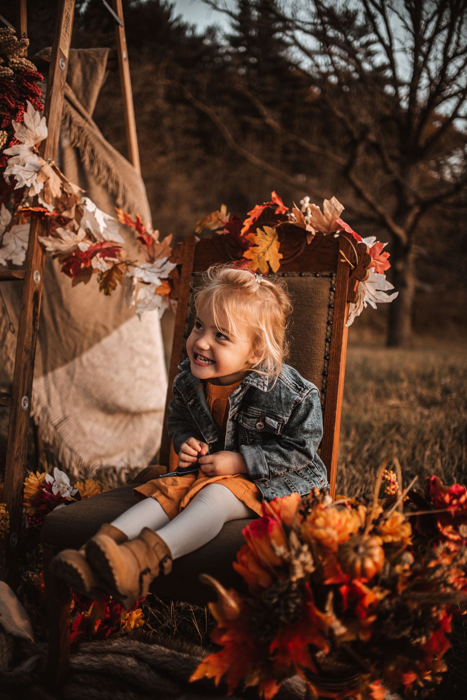 Autumn outdoor portrait of child in denim jacket sitting on vintage chair surrounded by fall leaves and decor.