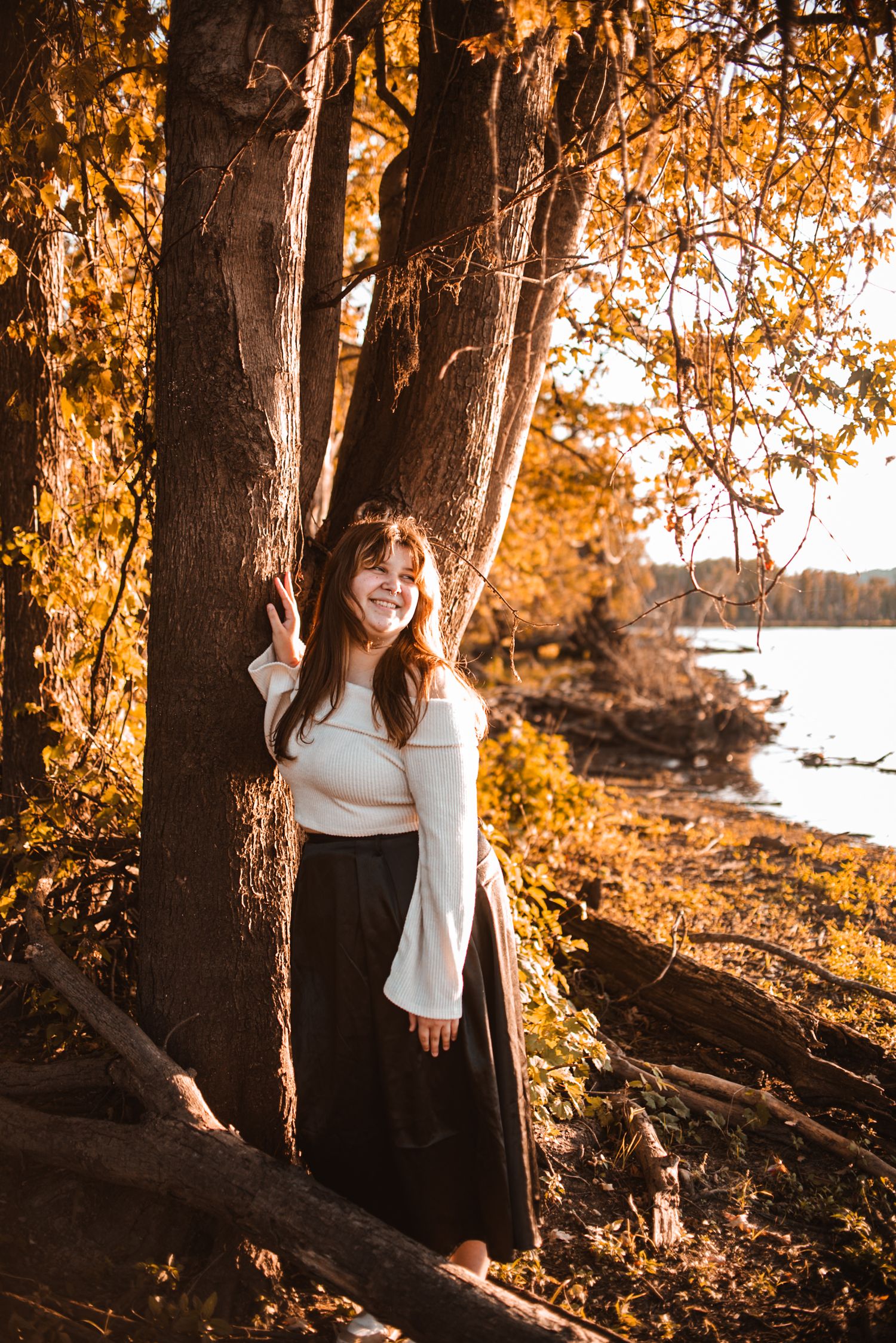 Person in white sweater and dark skirt stands by tree with autumn foliage reflecting in lake at sunset.