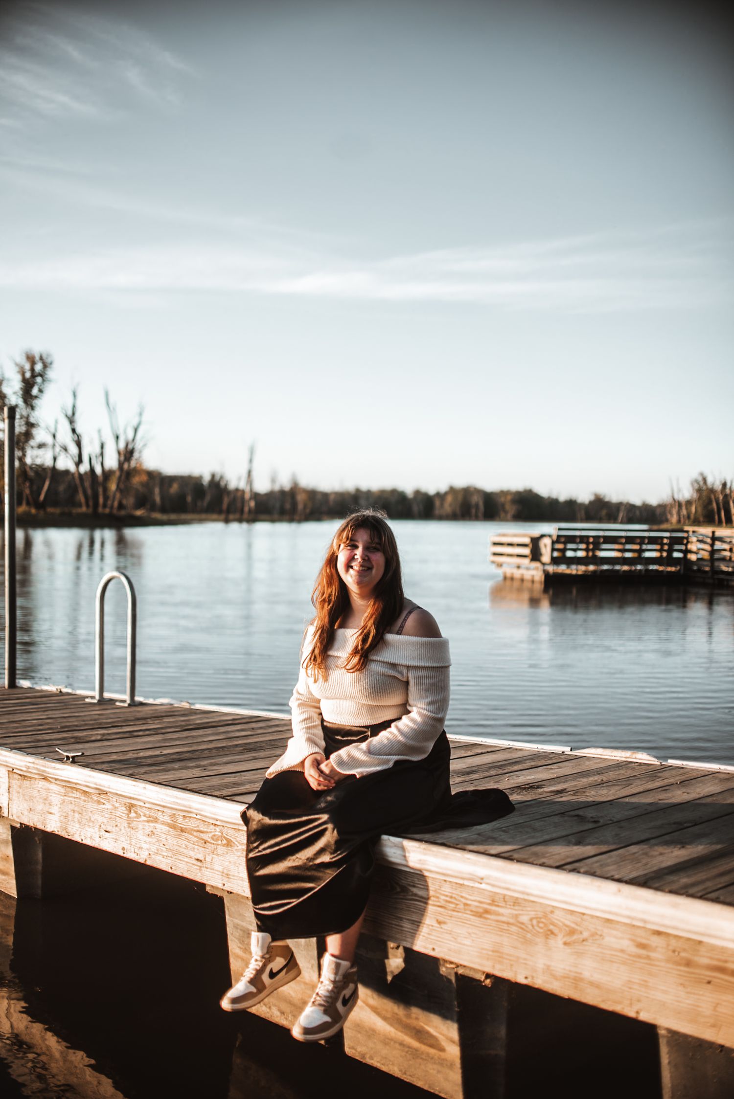 Someone sits on a wooden dock by a calm lake during golden hour with boats and trees visible in the background.