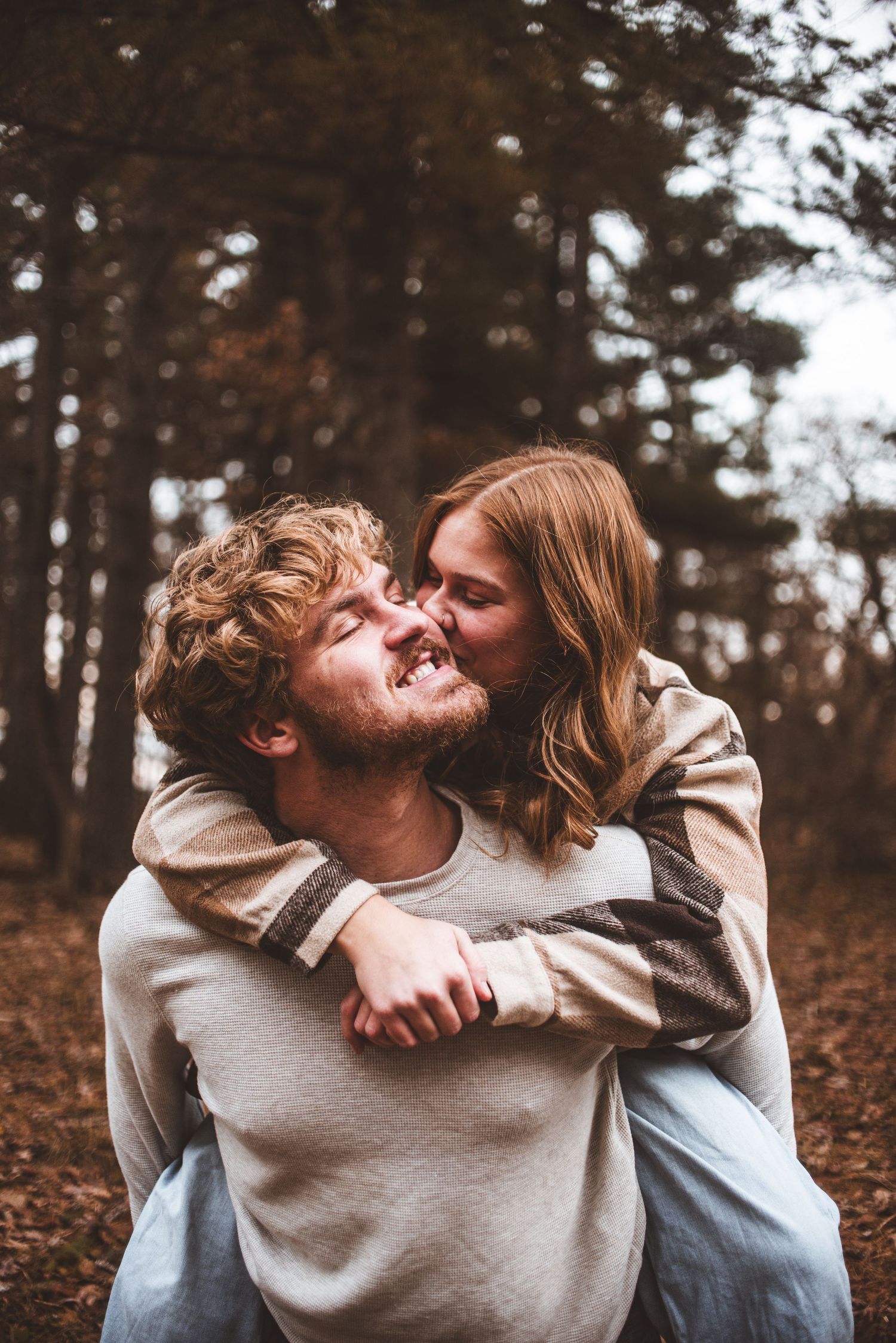 A couple shares a playful piggyback moment in a wooded forest setting on an autumn day.