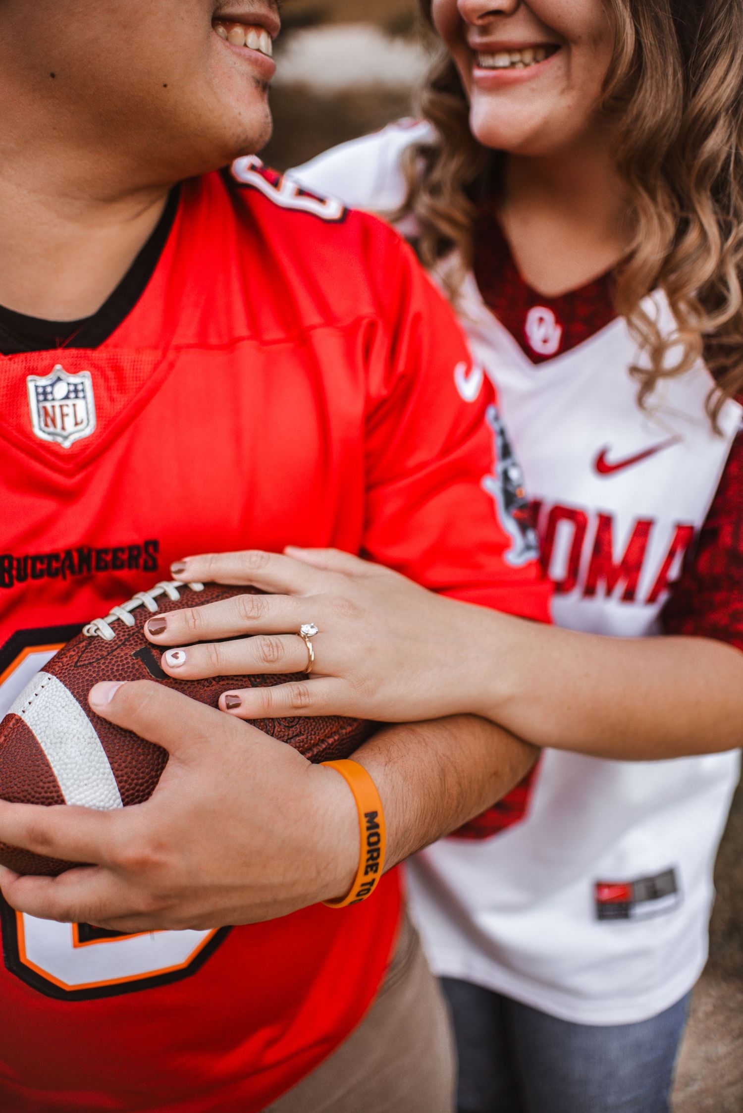 Engaged couple holds football together wearing NFL Buccaneers jersey and Oklahoma Sooners jersey while smiling.