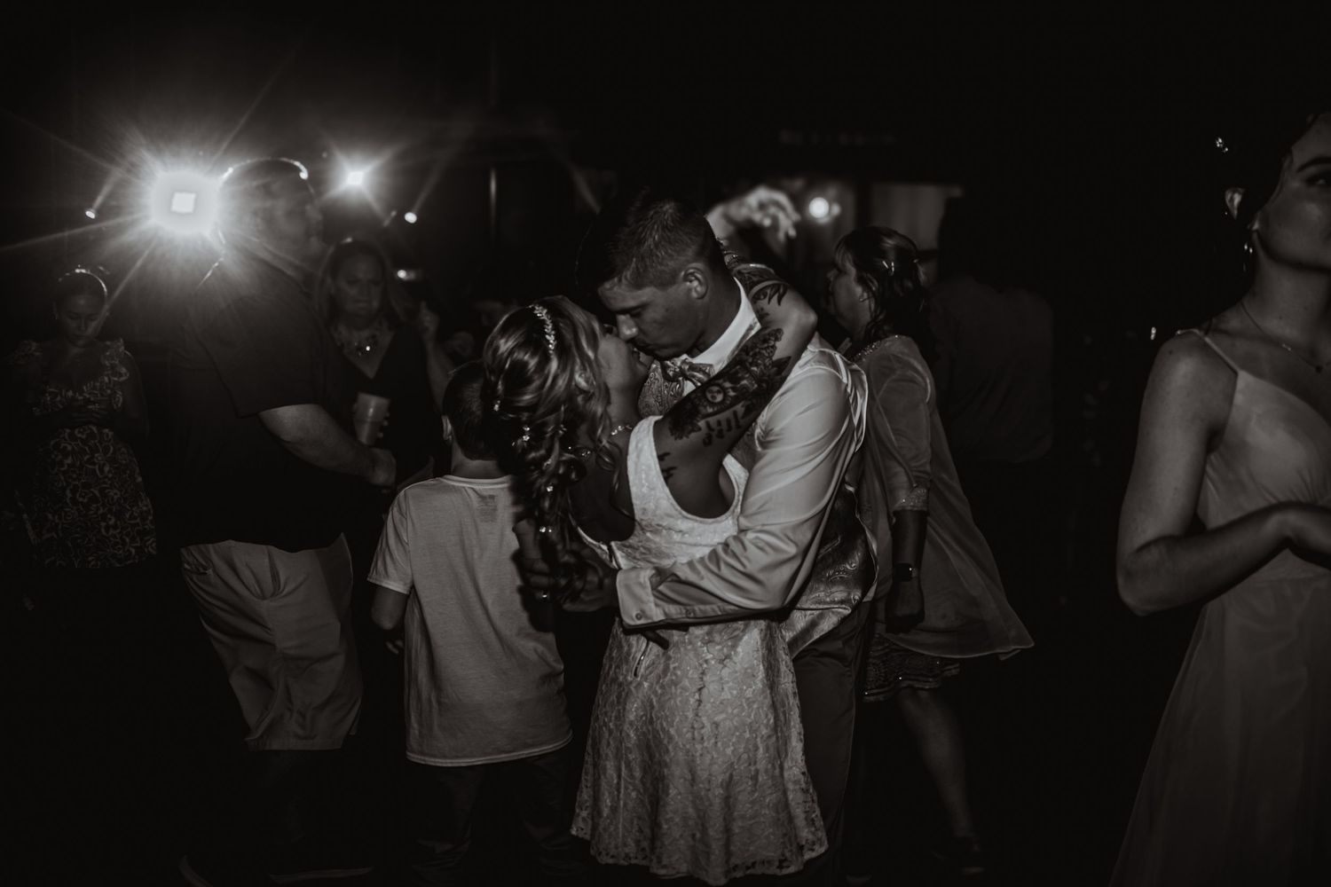 Dancing couple embracing at nighttime wedding reception with lights in background.