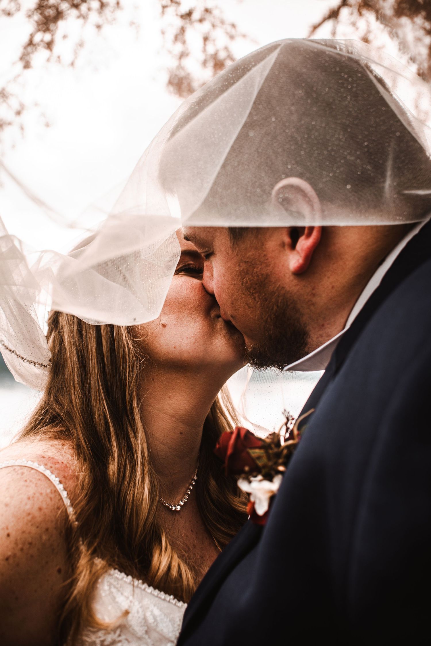 Romantic wedding photograph of a couple kissing under a flowing veil during their outdoor ceremony.