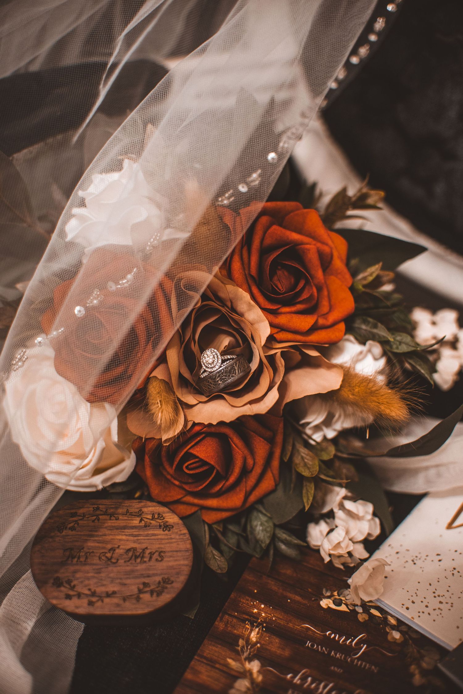 A romantic wedding detail shot with orange roses, sheer veil, and engagement ring on rustic wooden surface.