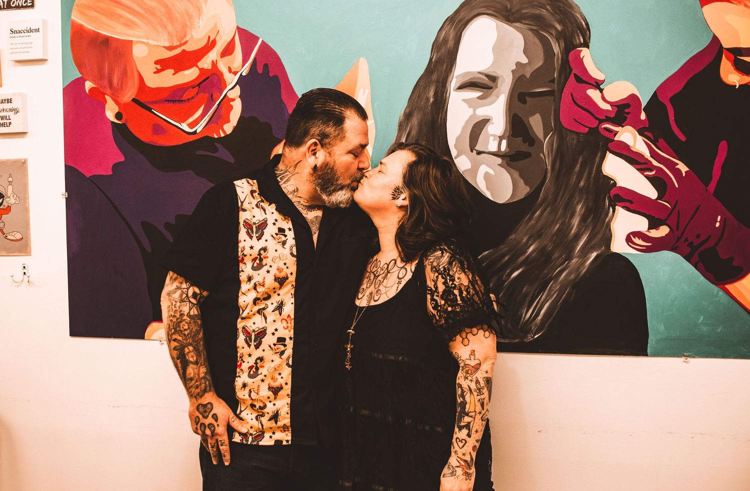 Two people stand together in front of a colorful artistic wall mural.