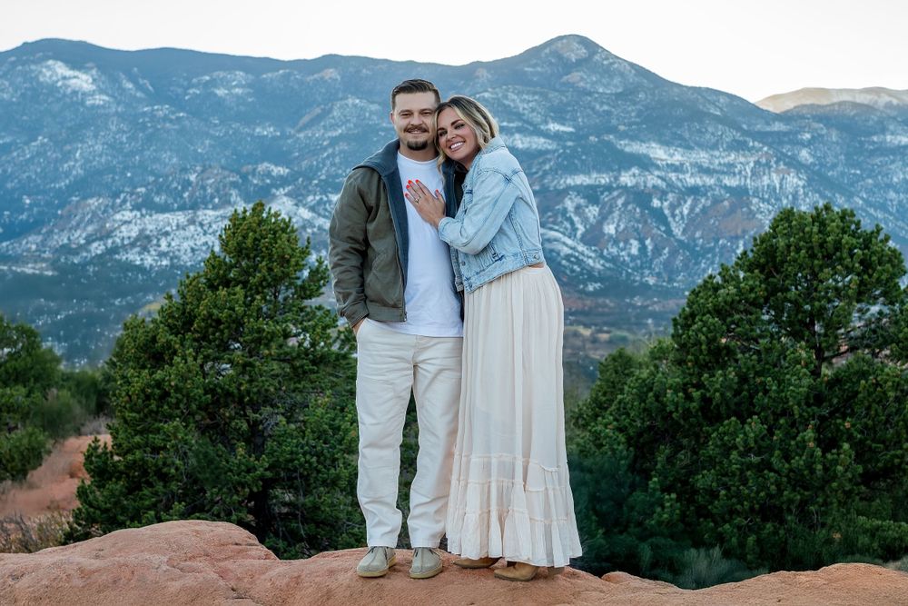 Couple embraces while standing on red rock formations with mountain vistas in the background.