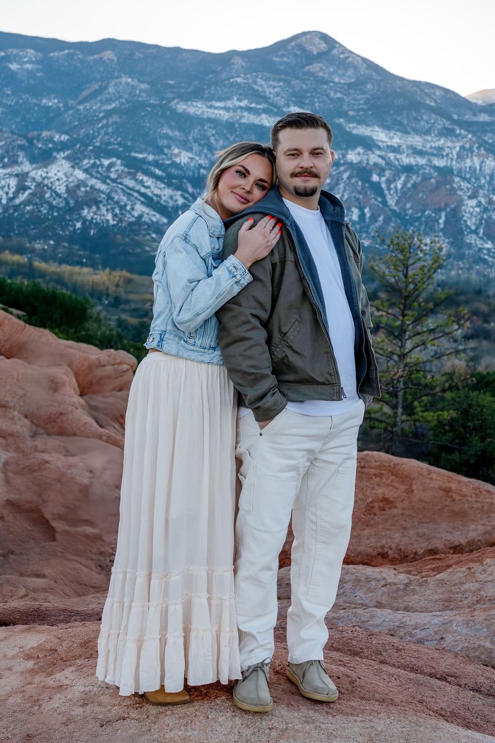 Couple in coordinated white and denim outfits pose on red rocks with mountain landscape backdrop.