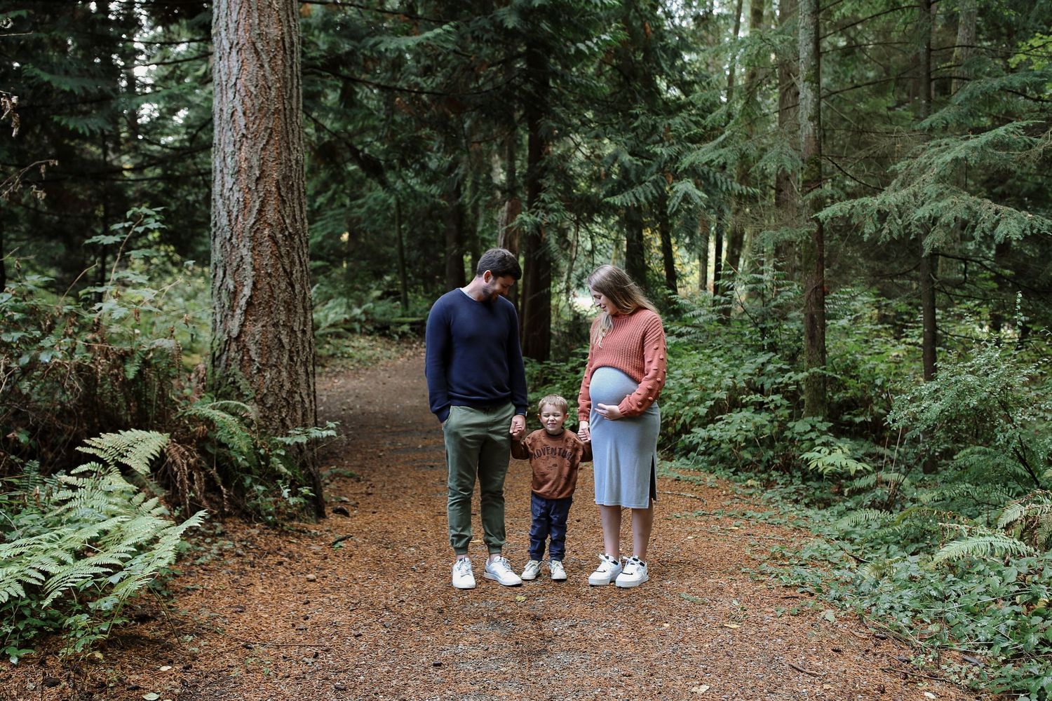 Family stands together on forest trail surrounded by tall evergreen trees.