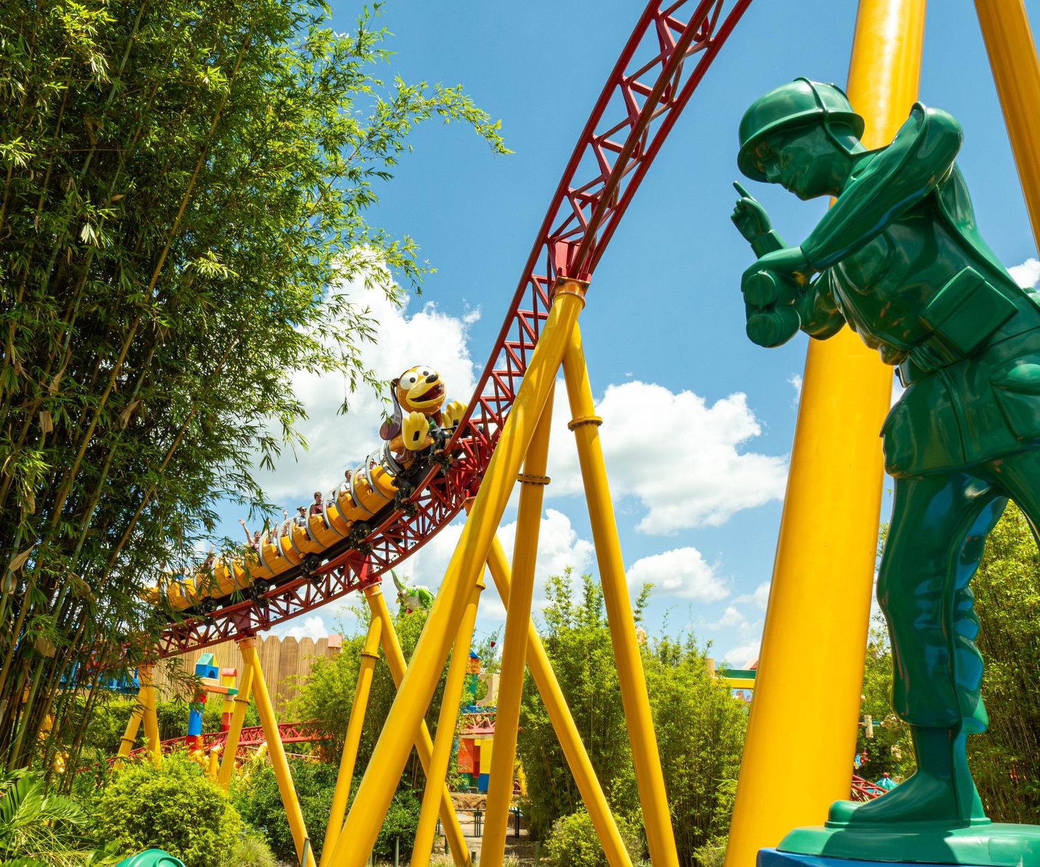 Bright yellow and orange roller coaster track curves around lush green trees against a vibrant blue summer sky.